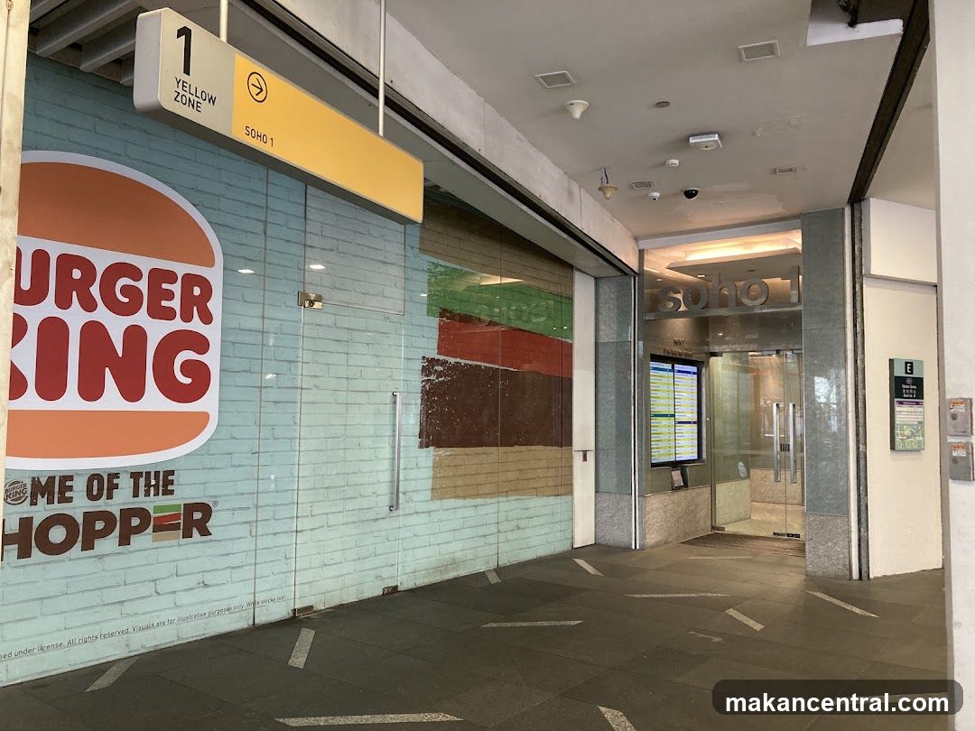 Exterior view of a Burger King store with a large logo on a light blue brick wall. Above the entrance is a yellow sign indicating 'Yellow Zone SOHO 1'. To the right, an adjacent glass door leads into a building labeled 'soho 1' with a digital display board inside.