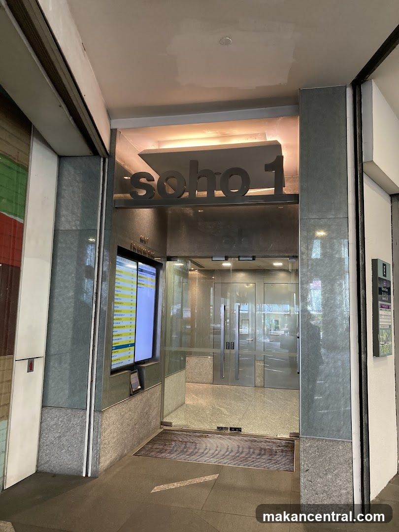 Modern glass entrance with 'soho 1' sign above the doorway. The facade features polished stone pillars and a digital display screen to the left showing a list of items or services. A metal grate is visible at the floor level leading into the lobby.