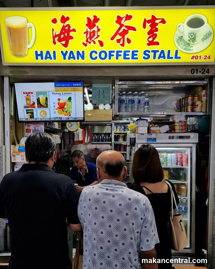 A storefront with a bright yellow sign featuring red Chinese characters "海燕茶室" and blue English text "HAI YAN COFFEE STALL #01-24". Customers are seen from behind at the counter, facing an employee inside. A refrigerator stocked with beverages is visible on the right. Behind the counter, there's a digital menu board showing drink options like Honey Lemon and Cold Brew. Water bottles and canned drinks are stored above. The shop number 01-24 is displayed on the right side of the sign.