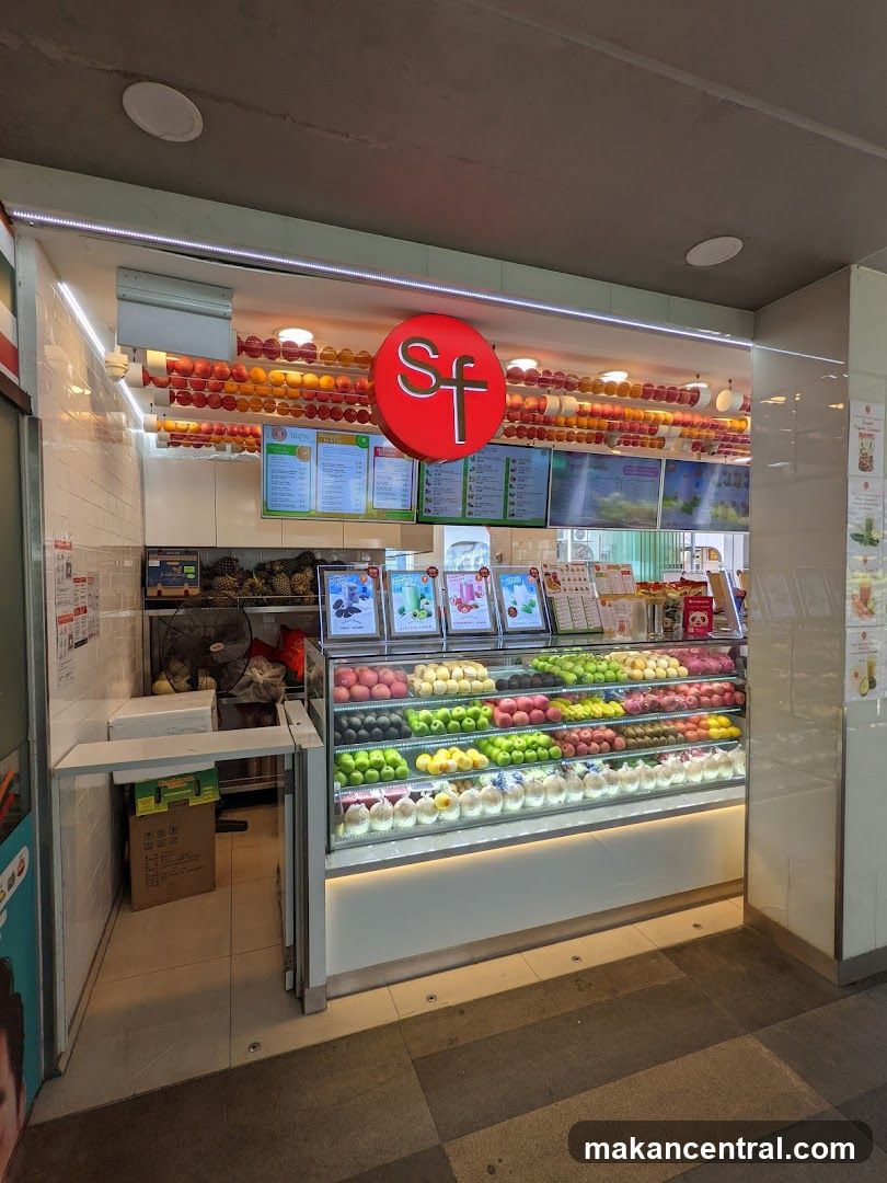 Storefront with illuminated red circular logo displaying 'Sf' above a refrigerated display case filled with assorted fruits. Above the counter, multiple digital screens show menus and promotional images. The shop has bright lighting and tiled walls. A fan is visible in the back area near stacked produce boxes.