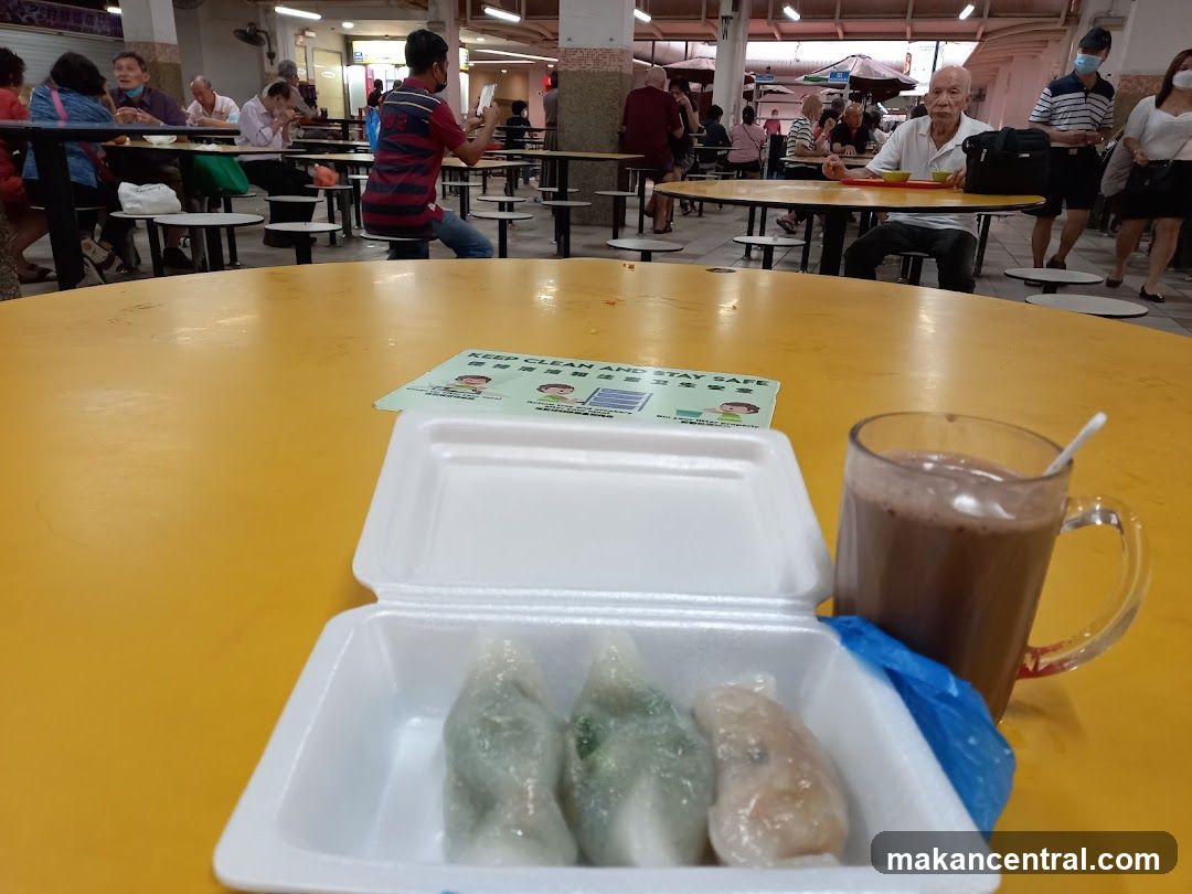 Yellow tables with food containers and a mug of chocolate drink in foreground. Background shows people dining at various tables, some wearing masks. A printed 'KEEP CLEAN AND STAY SAFE' poster is on the table. The setting appears to be an indoor hawker center or food court.