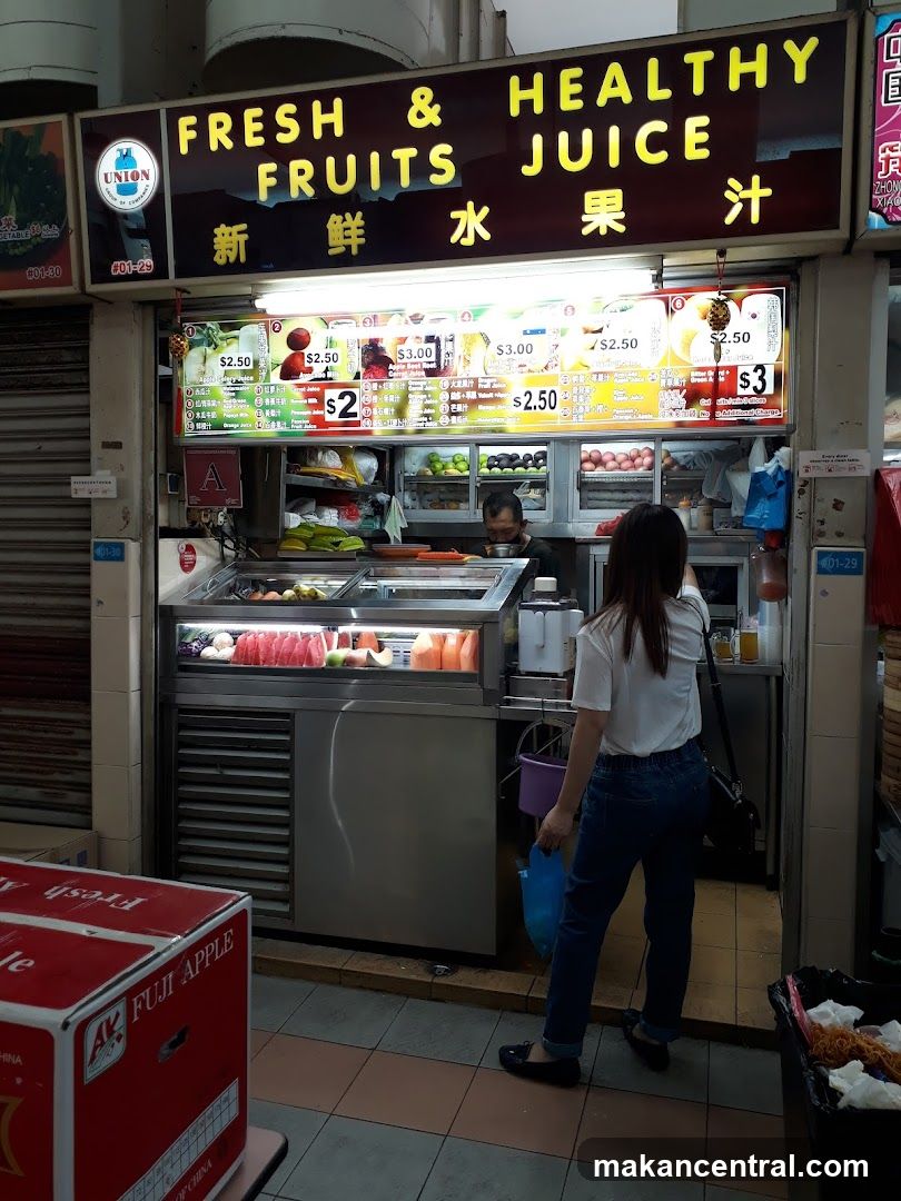 A storefront with a prominent illuminated sign reading 'FRESH & HEALTHY FRUITS JUICE' in English and Chinese characters. Below the main sign is a digital price display showing various juice options with prices like $2.50, $3.00, etc. The shop has refrigerated displays of fresh fruits and juicers inside. A customer stands at the counter while an employee works behind the counter. To the left, there are stacked red boxes labeled 'FUJI APPLE'.
