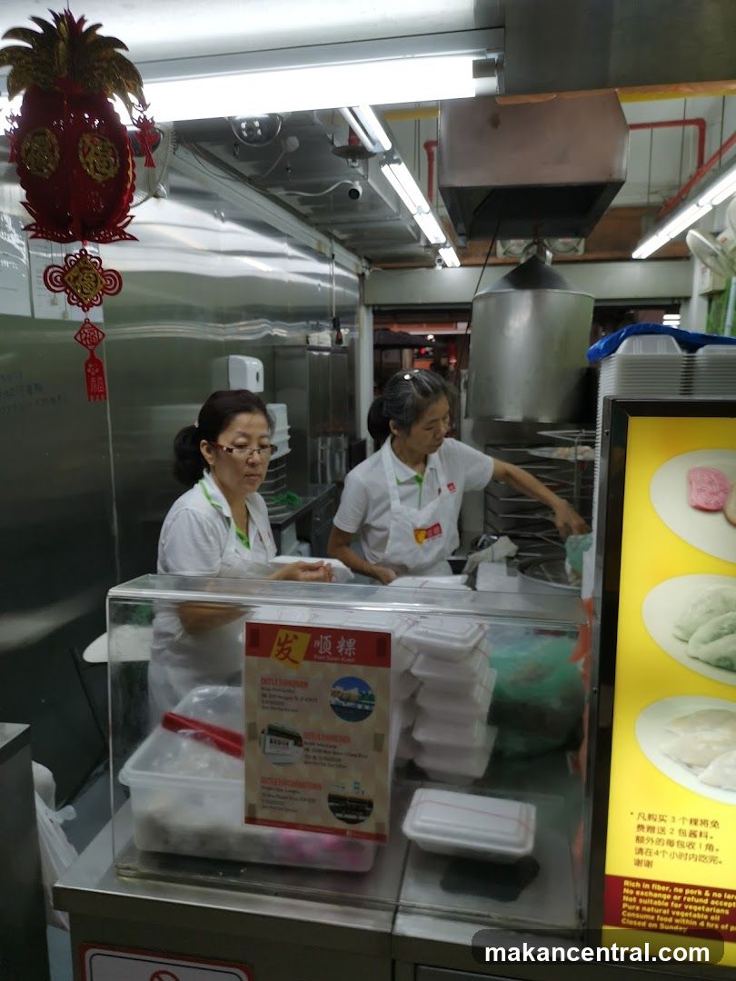 Two staff members in white uniforms are working behind a counter with food containers under glass display. A red decorative lantern hangs above. To the right is a yellow promotional board showing images of food items with Chinese text. The background shows stainless steel kitchen equipment and fluorescent lighting.
