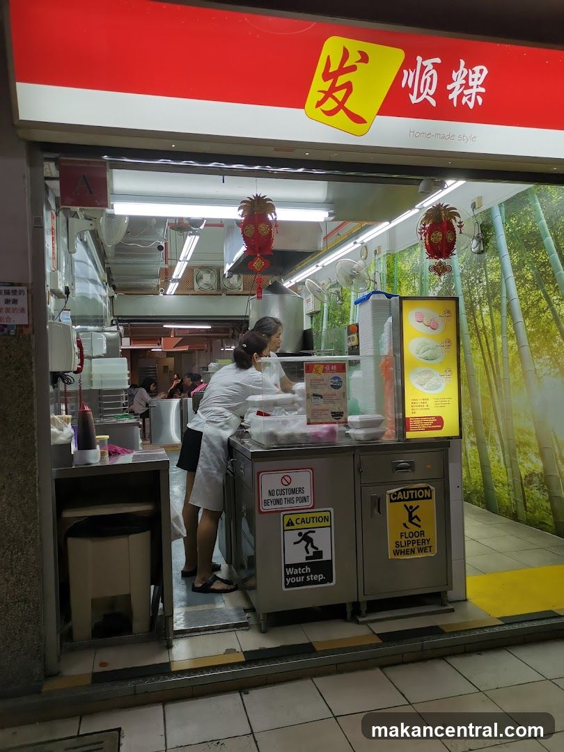 Exterior view of a food stall with a prominent red sign featuring yellow Chinese characters '发顺粿' and the phrase 'Home-made style'. Inside, staff in white uniforms are preparing food behind a counter. The area has safety signs including 'CAUTION FLOOR SLIPPERY WHEN WET' and 'NO CUSTOMERS BEYOND THIS POINT'. A bamboo-themed wallpaper decorates the right side wall. Two red lanterns hang from the ceiling.