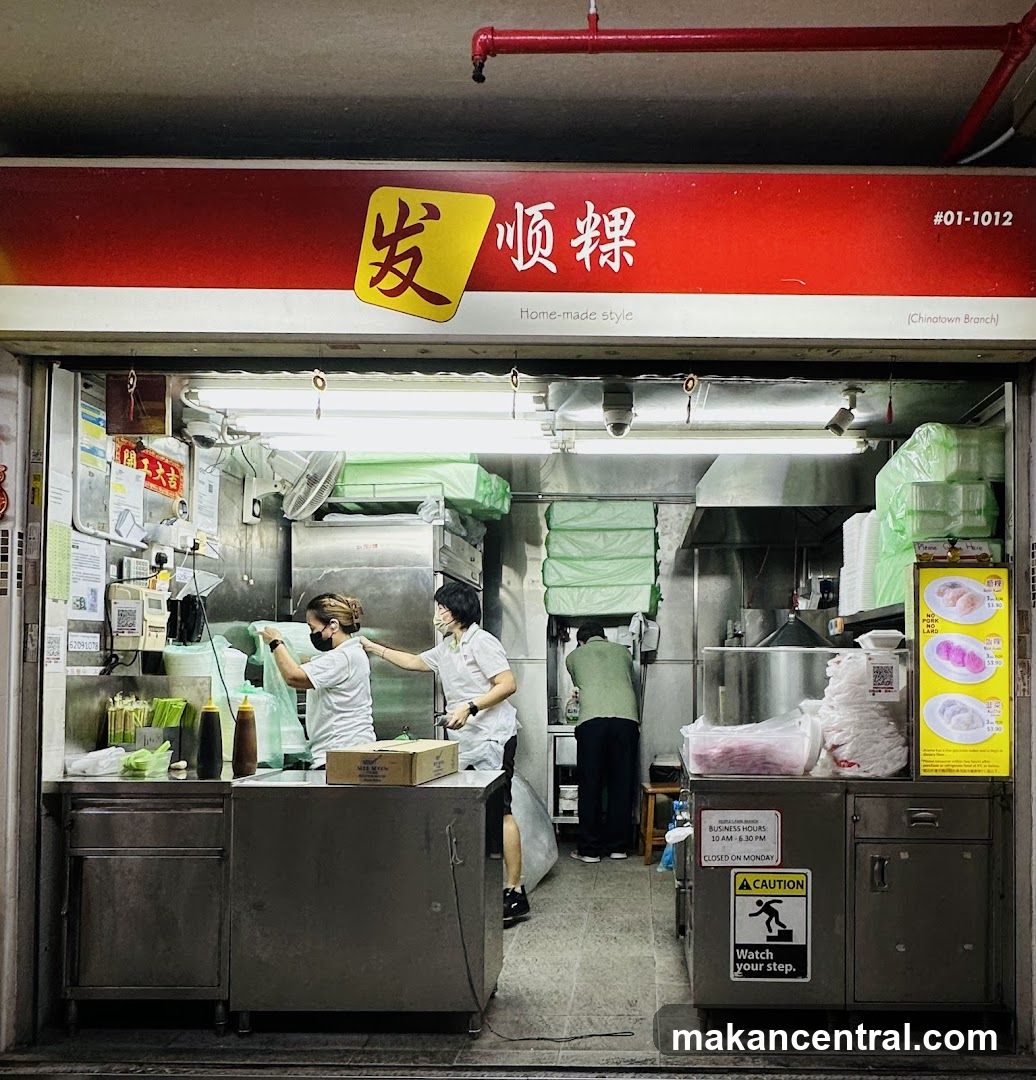 Exterior view of a food stall with red signage featuring Chinese characters '发顺粿' and the tagline 'Home-made style'. The shop is open with staff working inside, wearing masks and white uniforms. Stainless steel kitchen equipment and green-covered items are visible. A caution sign on the right cabinet reads 'Watch your step.' Above the entrance, a red pipe runs horizontally.