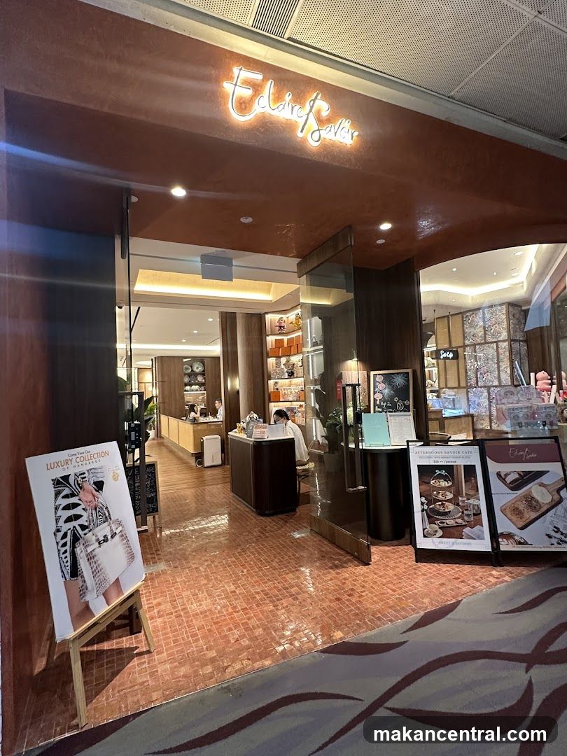 Storefront with illuminated neon sign reading 'Eclaire Savoir' above entrance. Glass doors lead into a warmly lit interior with wooden floors and display shelves. Two promotional stands outside feature branded merchandise and café offerings.