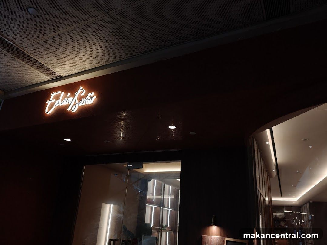 Nighttime photo of a modern restaurant entrance with illuminated cursive signage reading 'Elaine Savoir' mounted on a dark facade above glass doors. Interior lighting is visible through the doorway, showing contemporary design elements including vertical light strips and wooden accents.