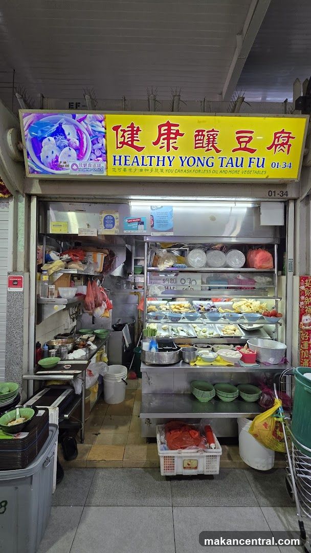 A storefront with a bright yellow sign featuring Chinese characters '健康醤豆腐' and English text 'HEALTHY YONG TAU FU 01-34'. The shop is open, revealing an interior with stainless steel counters, shelves stocked with food items, and cooking equipment. A digital display shows images of the dish. The entrance area includes stacked bowls, plastic bins, and a shopping cart.