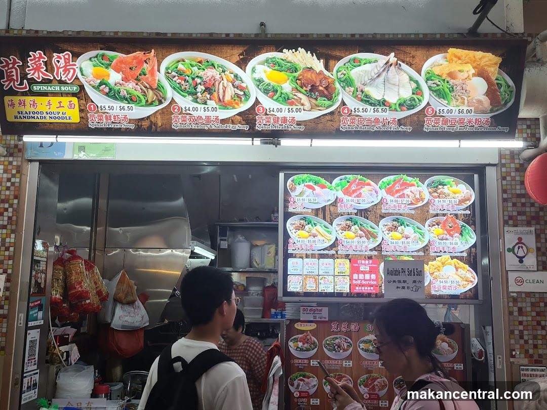 A food stall with a large overhead sign displaying five types of spinach-based dishes with prices. The interior is partially visible, showing stainless steel kitchen equipment and customers at the counter. A digital menu board displays additional options below the main sign.