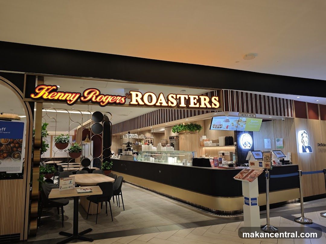 Storefront with illuminated sign above counter, modern interior featuring wooden accents, glass display cases, seating area with tables and chairs, order station with digital screens, and decorative plants.