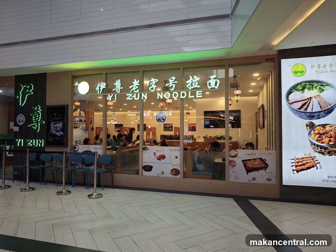 Exterior storefront of a noodle restaurant with large glass windows displaying interior seating and diners. Neon green signage reads '伊尊老字号拉面' and 'YI ZUN NOODLE'. A vertical neon sign to the left displays '伊尊 YI ZUN'. The floor is tiled in herringbone pattern, and there are blue chairs outside with stanchions.