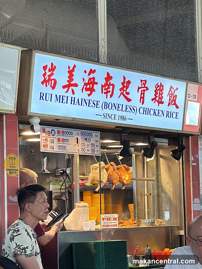 Exterior storefront with a large blue sign featuring red Chinese characters and English text 'RUI MEI HAINESSE (BONELESS) CHICKEN RICE — SINCE 1986'. Below the sign, a glass display case shows roasted chickens hanging above yellow rice bowls. A customer is seen at the counter holding a phone. The shop has a metal mesh screen above the entrance.