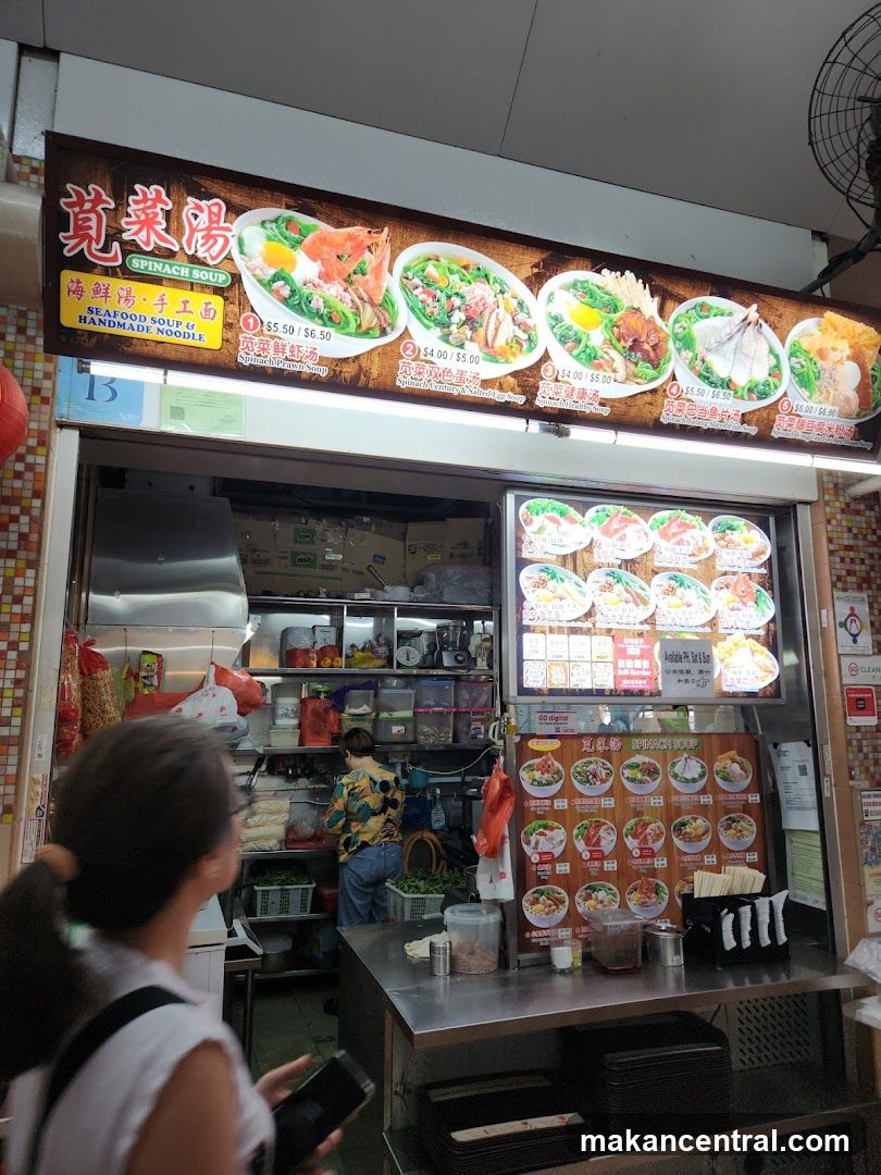 A food stall with a large signboard above the counter displaying images of dishes and prices in Chinese and English. The interior shows a kitchen area with shelves, appliances, and a staff member working. A customer is seen from behind at the counter. The storefront has a tiled wall and a fan mounted on the ceiling.