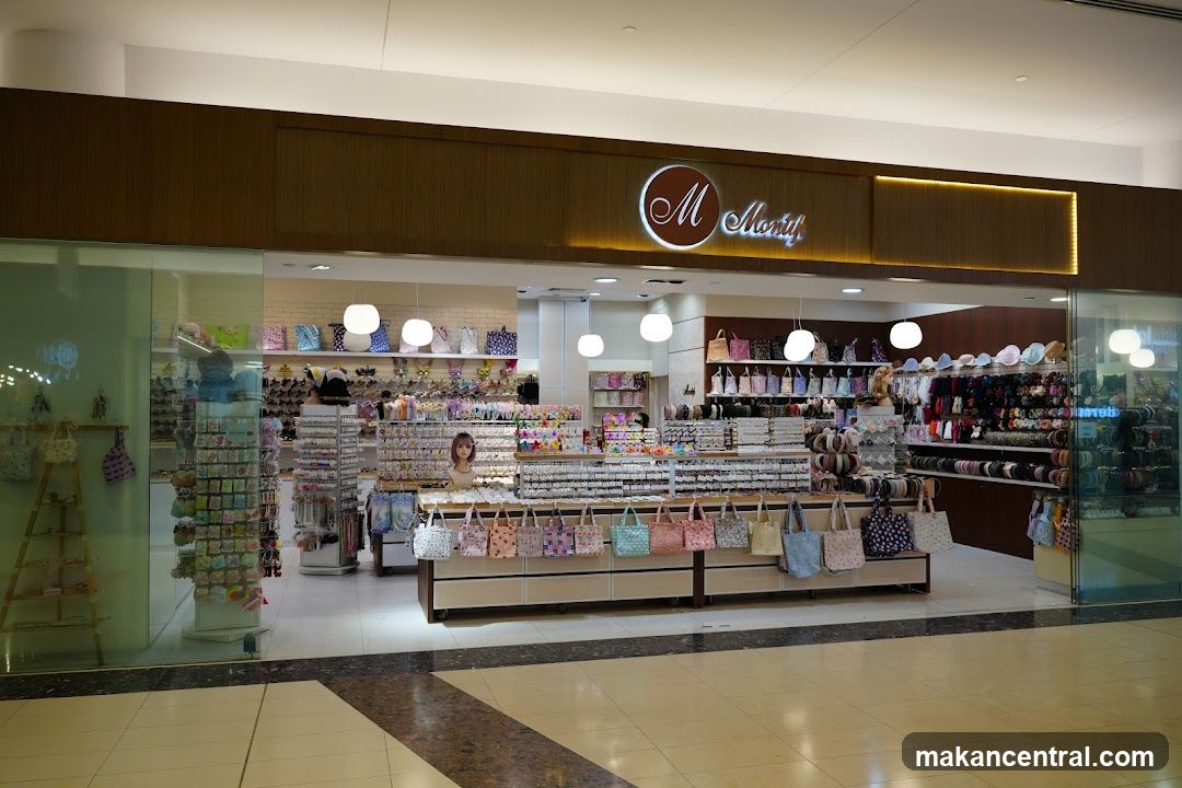 Storefront with illuminated logo 'M Manifi' on a wooden panel above glass display windows. Interior shows shelves stocked with bags, ribbons, and accessories under bright lighting. A counter displays patterned handbags in front. No menu or printed materials are visible.