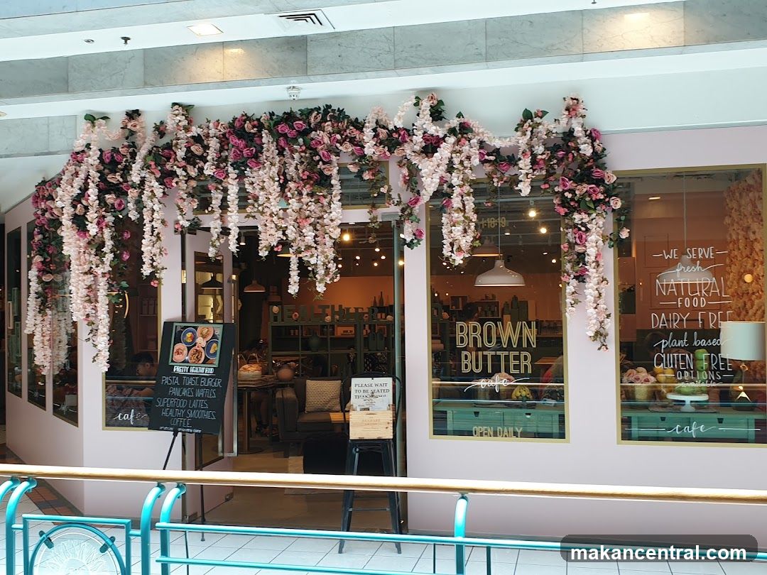 Storefront facade with pink and white floral garlands hanging above the entrance. Large glass windows display signage: 'BROWN BUTTER cafe', 'OPEN DAILY', and 'WE SERVE NATURAL FOOD DAIRY FREE plant based GLUTEN FREE OPTIONS'. A chalkboard menu stand is positioned outside, but not fully legible. Interior lighting and seating are partially visible through the glass.