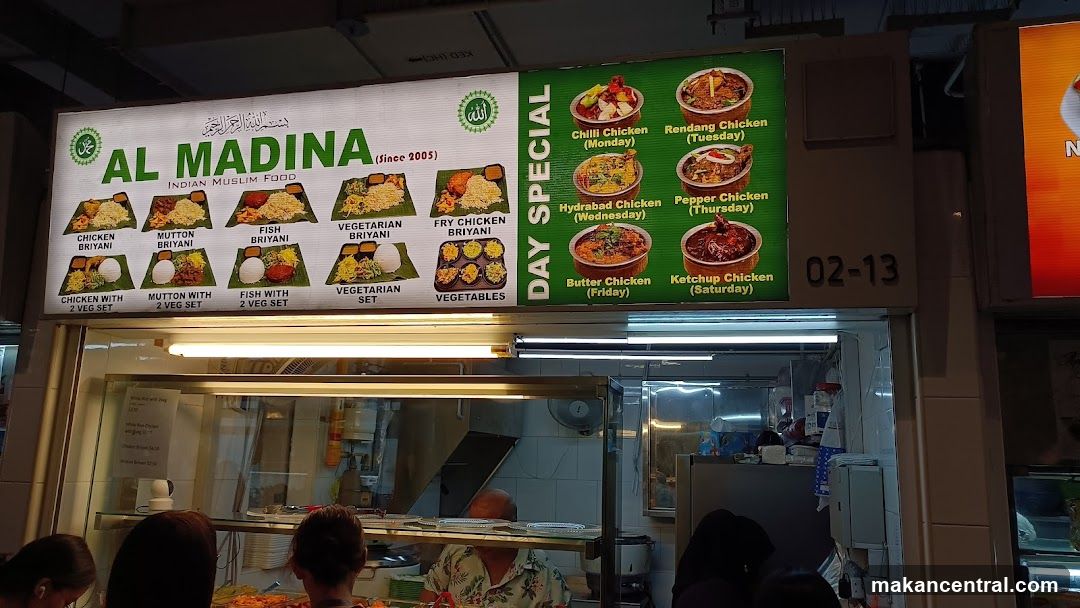 Exterior storefront of a food stall with illuminated signage above the counter. The sign displays 'AL MADINA' and 'INDIAN MUSLIM FOOD (since 2005)'. Below, images and names of dishes are shown under categories like 'CHICKEN BRIYANI', 'MUTTON BRIYANI', etc., alongside 'DAY SPECIAL' featuring daily chicken dishes. A staff member is visible behind the counter. The stall number '02-13' is displayed on the right.
