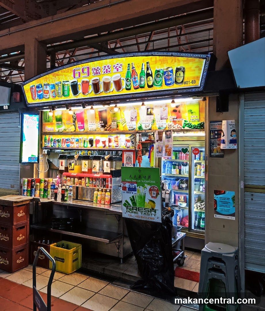 Brightly lit storefront with a yellow sign displaying beverages and the number #01-69. The shop is open with visible drink dispensers, refrigerated units, and stacked branded crates outside. A black plastic bag covers part of the entrance. To the right, a shuttered adjacent stall is partially visible.