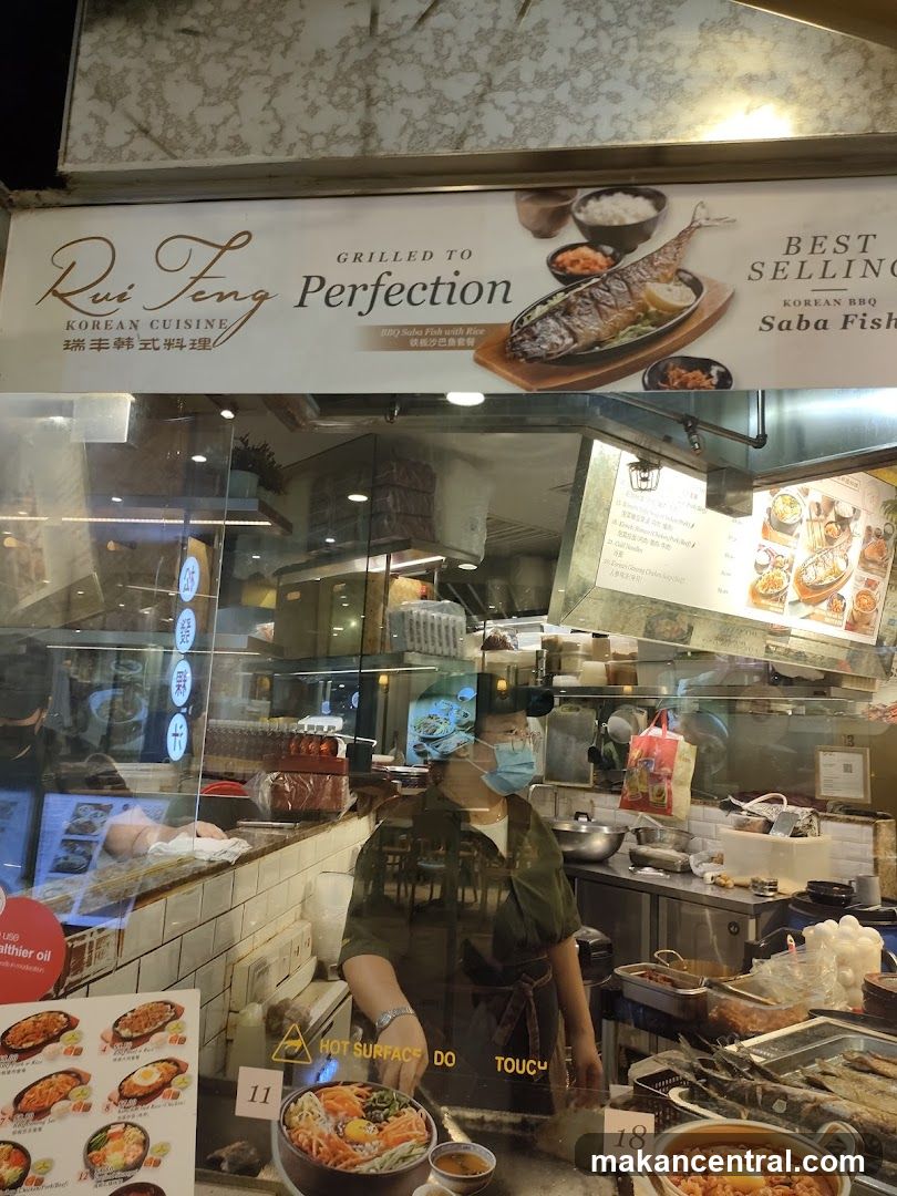 Storefront with glass window showing interior kitchen area; staff wearing mask working behind counter; sign above reads 'Rui Feng Korean Cuisine' and 'Grilled to Perfection', featuring image of grilled fish on plate. Sign also includes 'Best Selling Korean BBQ Saba Fish'. Reflections and interior lighting visible.