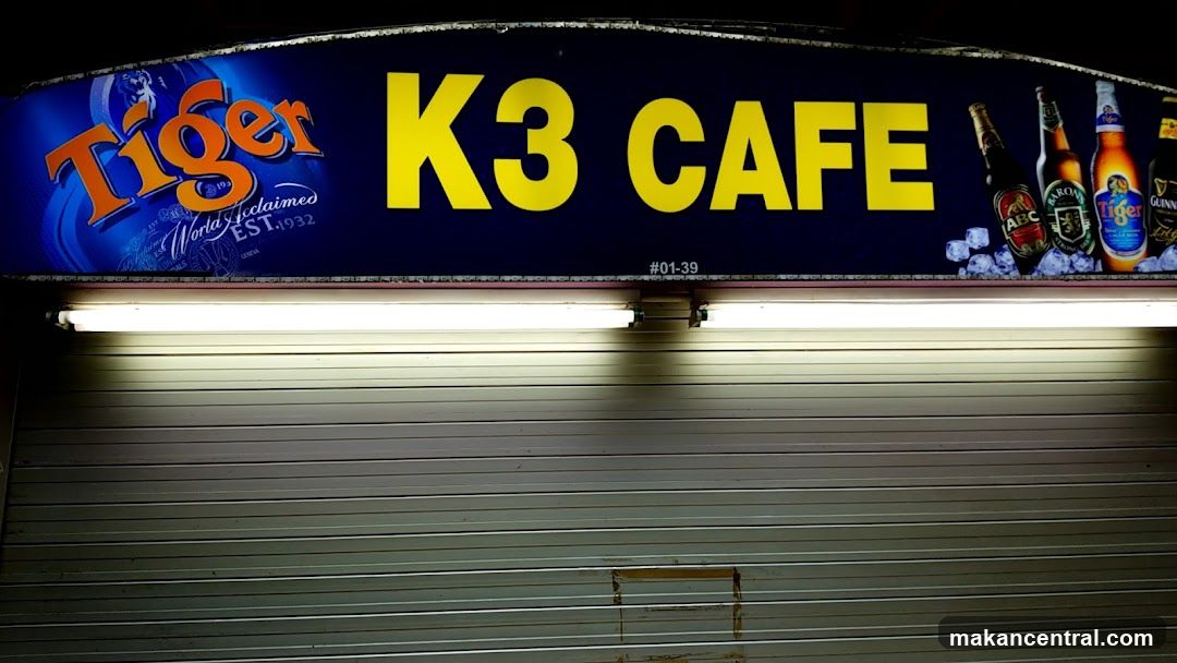 Nighttime exterior shot of a closed storefront with a bright blue sign featuring 'Tiger' logo on the left and 'K3 CAFE' in large yellow letters center-right. The sign also displays various beer brands (ABC, Guinness, Tiger) on ice cubes to the right. Below the sign is a closed metal roll-down shutter door. A fluorescent light fixture runs horizontally beneath the sign. Unit number #01-39 is visible below the main text.
