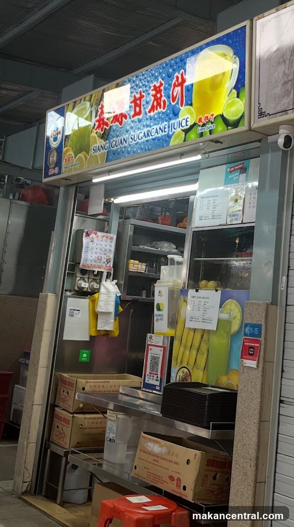Exterior storefront of a sugarcane juice stall with illuminated signboard featuring Chinese characters '泰源甘蔗汁' and English text 'SIANG GUAN SUGARCANE JUICE'. The shop has a glass window revealing interior equipment, stacked cardboard boxes, red plastic stools, and QR code payment stickers on the wall. A security camera is mounted above right.