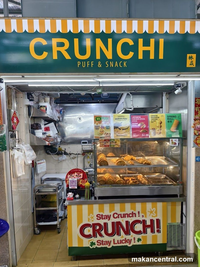 A food stall with a green and yellow striped awning displaying 'CRUNCHI PUFF & SNACK' in large yellow letters. Below the counter, a sign reads 'Stay Crunch! Stay Lucky!' The interior shows a glass display case filled with fried snacks, stainless steel kitchen equipment, and promotional posters on the wall. A Chinese character is visible on the right side of the main sign.