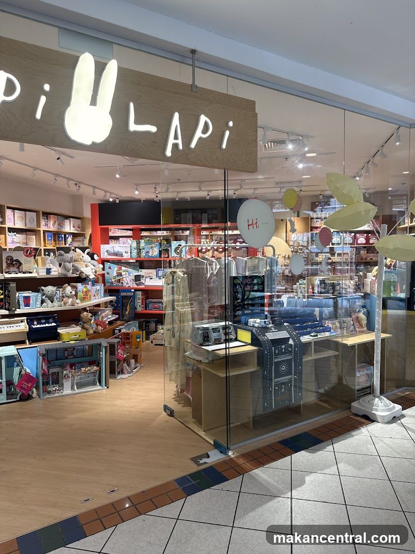 Storefront with wooden sign featuring illuminated 'LApi' logo and a stylized rabbit silhouette. Glass display windows show interior shelves stocked with toys and books. A transparent barrier separates the store from the walkway. Decorative balloons hang near the entrance, including one labeled 'Hi...'. The floor features tiled edges with multicolored tiles.