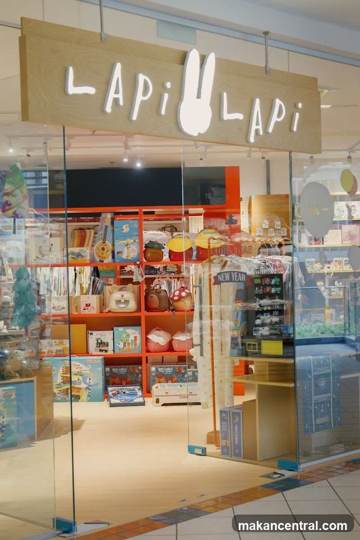 Glass storefront with wooden sign displaying 'LApi LApi' in white letters, featuring a bunny silhouette. Inside, orange shelving units display toys, books, and seasonal items like 'New Year' decor. The shop is brightly lit with overhead lighting.