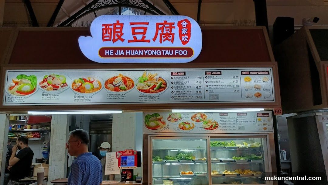 A food stall with a bright illuminated sign displaying the name in Chinese characters (酿豆腐) and English (HE JIA HUAN YONG TAU FOO). The sign is mounted above the storefront, which includes a glass display case filled with fresh vegetables and prepared dishes. Customers are visible inside the stall area.