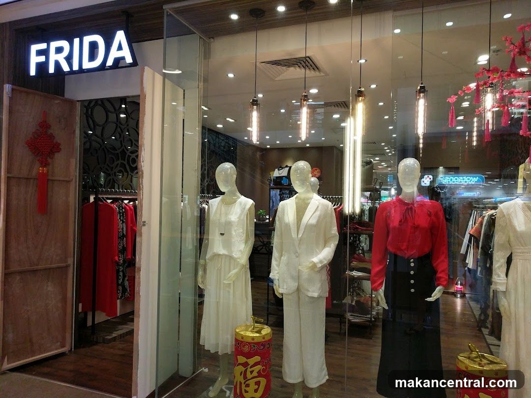 Store entrance with illuminated "FRIDA" sign above glass door. Display window features mannequins in white and red attire. Red Chinese knot decor hangs on wooden panel to the left. Interior lighting and hanging lamps are visible through glass. Red decorative lanterns adorn ceiling right side.