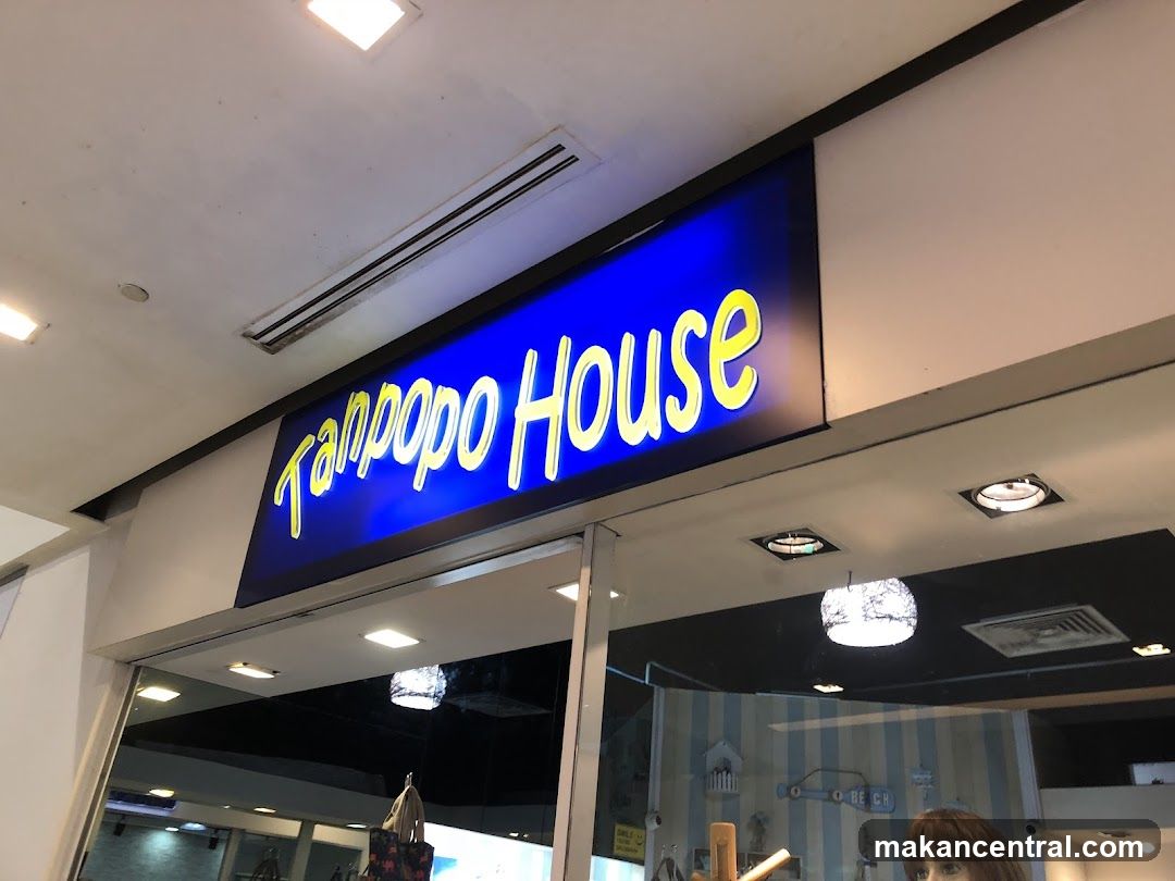 A brightly lit blue sign with yellow lettering reading 'Tampopo House' is mounted above a glass entrance door. The interior is partially visible through the glass, showing striped wallpaper and hanging lights. The ceiling has recessed lighting and ventilation grilles.