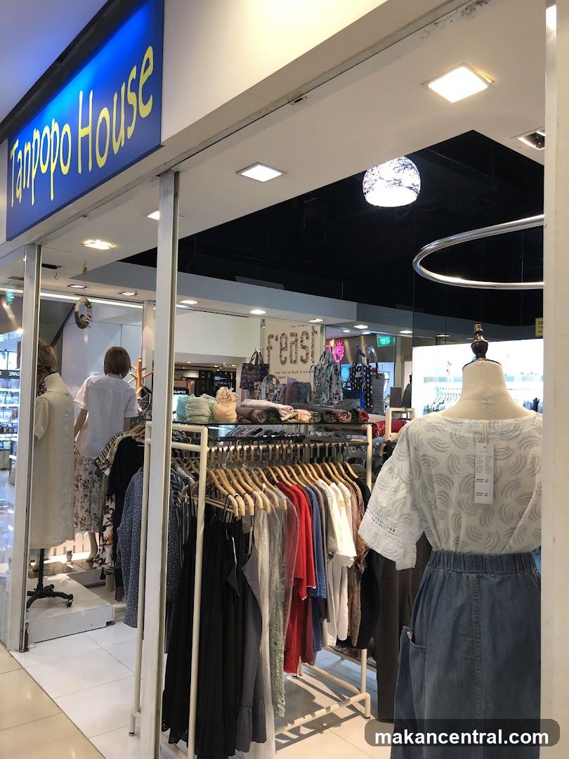Storefront with blue sign bearing 'Tanpopo House' in yellow lettering. View from outside or doorway looking into the shop interior showing racks of clothing, mannequins, and a display shelf with folded garments. Interior lighting includes ceiling lights and a decorative spherical pendant light. A partial view of another store's signage ('FEAST') is visible in background.