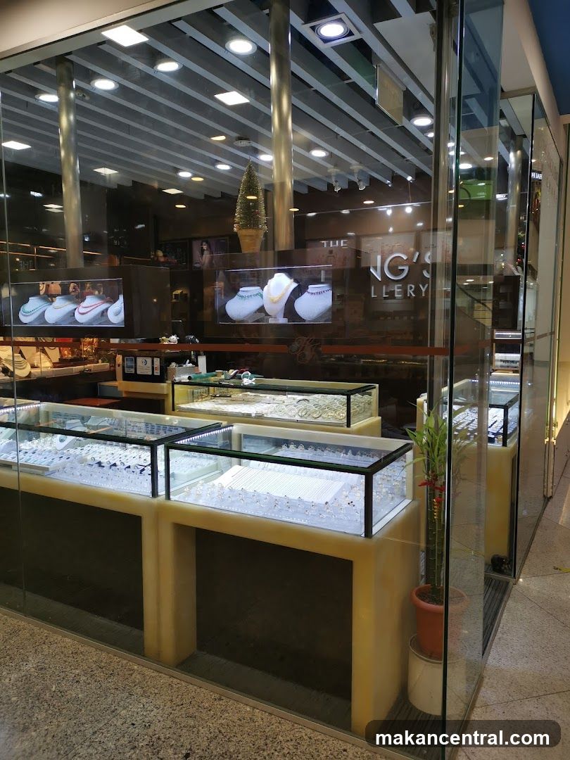Glass storefront with illuminated display cases showcasing jewelry. A small potted plant stands beside the entrance. Reflections show interior lighting and a Christmas tree in the background. Signage on glass reads 'THE GALLERY' partially obscured by reflections.