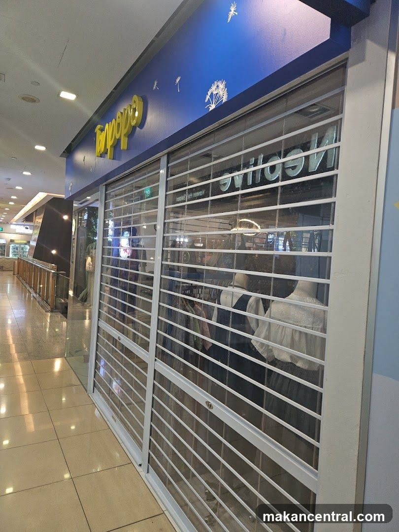 A storefront with blue signage featuring yellow lettering above a set of closed white roll-down metal security doors. The glass panels behind the doors show mannequins dressed in clothing. Reflections of the interior lighting are visible on the glass. The floor is tiled, and the ceiling has recessed lights.