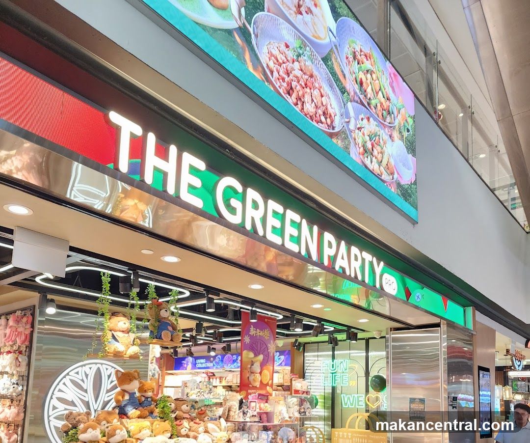 Storefront with illuminated sign reading 'THE GREEN PARTY PRO' in white letters on a green background. Above the sign is a large digital display showing food dishes. The entrance features stuffed animals and decorative elements inside the shop. Glass doors and neon signs are visible, including one that says 'WE ❤️'.