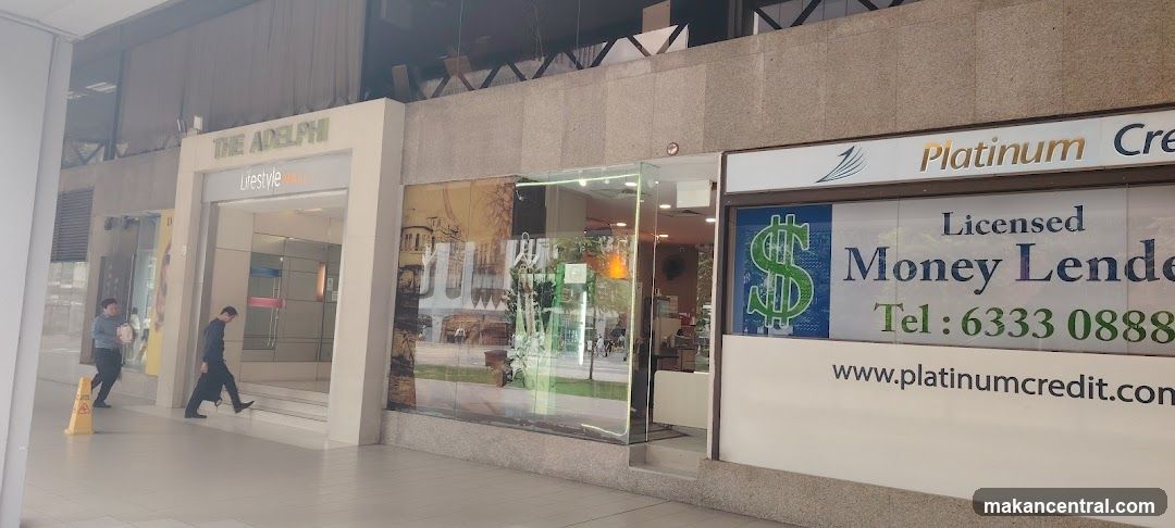 Exterior view of a commercial building facade with two adjacent storefronts. On the left, 'THE ADELPHI' sign above an entrance labeled 'Lifestyle'. To the right, a glass-fronted shop with signage for 'Platinum Credit', featuring a large green dollar sign logo and contact details including phone number 6333 0888 and website www.platinumcredit.com. Two pedestrians are visible walking past the entrance.
