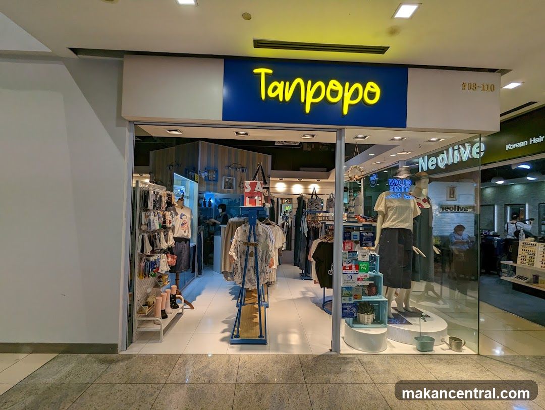 Store entrance with bright yellow neon sign reading 'Tanpopo' on a blue background. Glass storefront displays mannequins wearing clothing, racks of garments inside, and promotional signage including a red 'SALE' sign. Adjacent store signs show 'Neolive' and 'Korean Hair'. The floor is tiled, ceiling has recessed lighting.