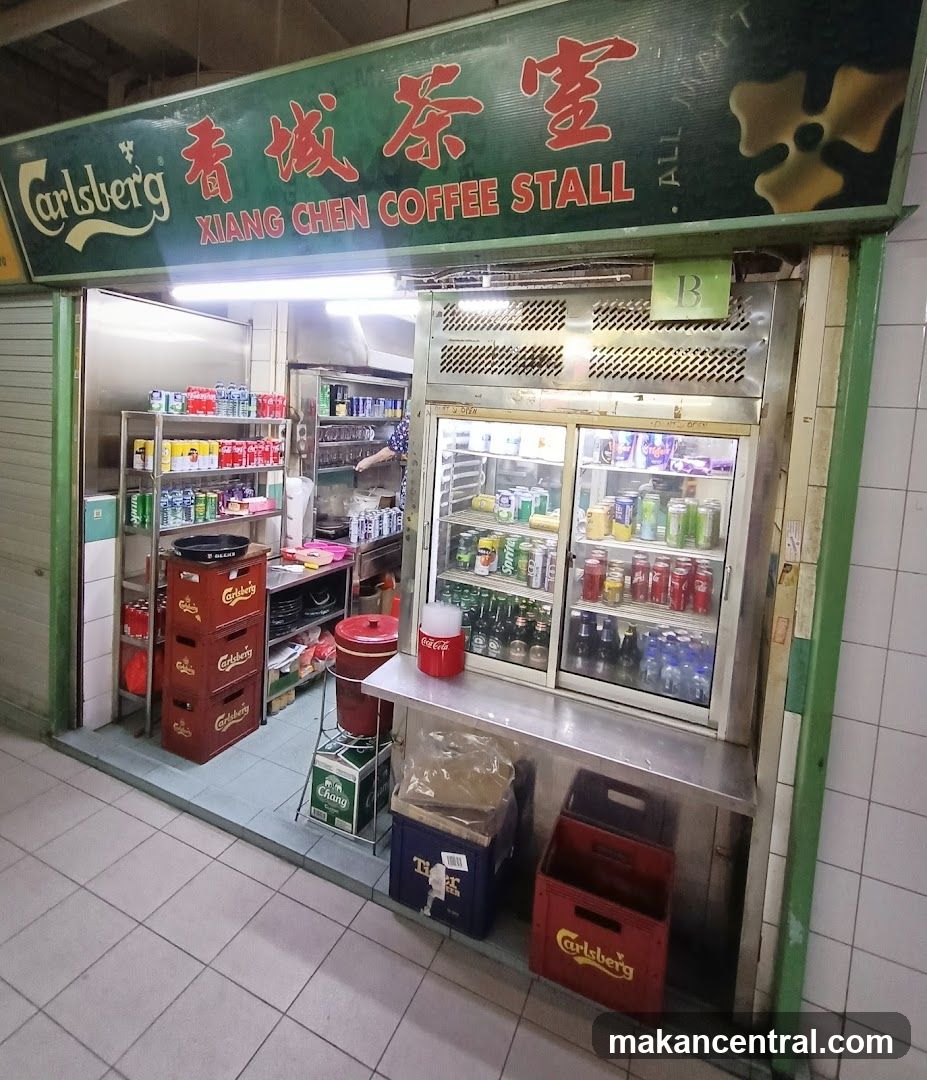 A storefront with a green sign featuring red Chinese characters "香城茶室" and English text "XIANG CHEN COFFEE STALL". The sign also includes the Carlsberg logo on the left. Inside, there are refrigerated display units filled with beverages, shelves stocked with canned drinks, and stacked branded crates (Carlsberg, Chang) outside. A person is partially visible inside preparing food or drink.