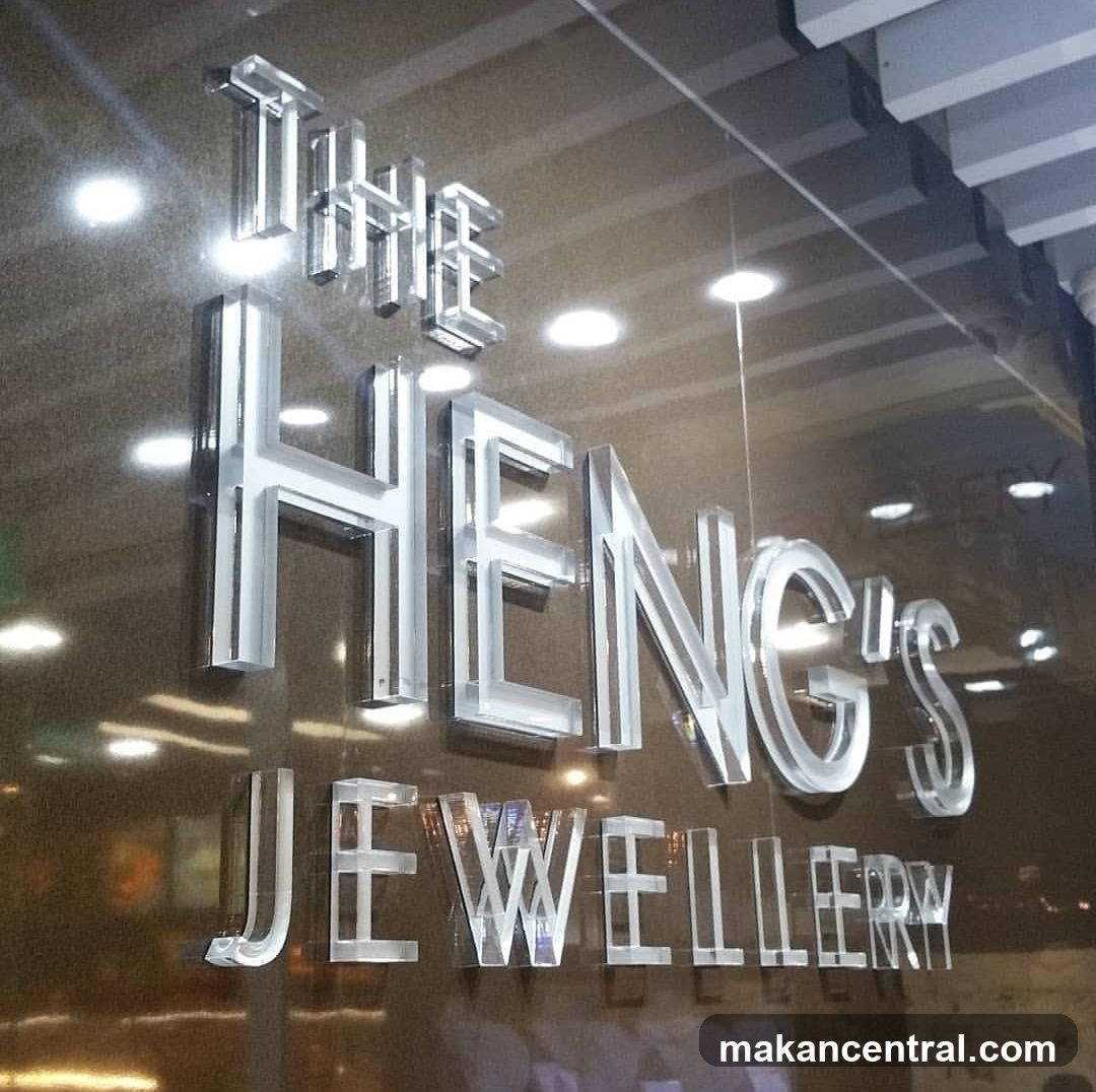 A storefront with large, illuminated silver 3D lettering on a reflective glass surface. The sign reads 'THE HENG'S JEWELLERY'. Reflections of interior lights and ceiling fixtures are visible on the glass. No other details like doors or windows are clearly shown.