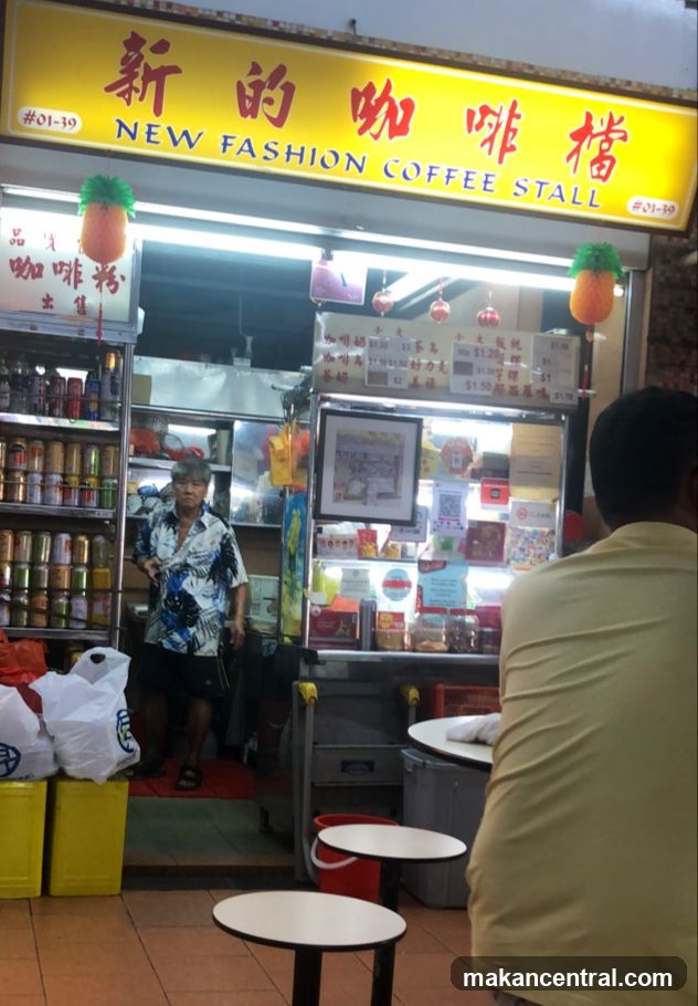 Exterior storefront with bright yellow signboard displaying "新的咖啡檔" and "NEW FASHION COFFEE STALL" in red and black lettering. Two pineapple-shaped lanterns hang from the ceiling. A staff member stands inside near shelves stocked with canned goods. Customers are seated at small round tables outside. The shop entrance is open, revealing interior lighting and a counter area.