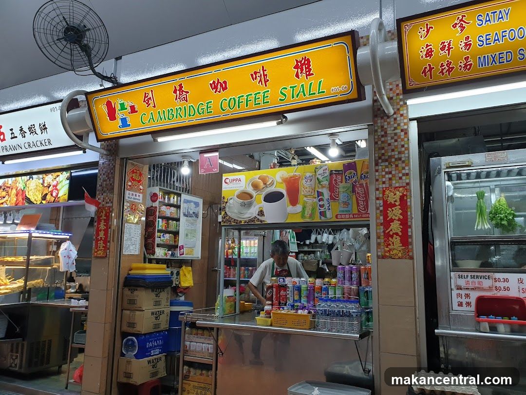 A storefront with a bright yellow sign featuring red Chinese characters and green English text "CAMBRIDGE COFFEE STALL" above the entrance. The shop has a glass window display showing beverages and snacks inside. A staff member is visible behind the counter, surrounded by canned drinks and bottled water. To the right, another stall's sign reads "沙嗲 SATAY 海鮮湯 SEAFOOD 什锦汤 MIXED S". Above, a ceiling fan hangs. The interior shows shelves stocked with goods and promotional posters on the walls.
