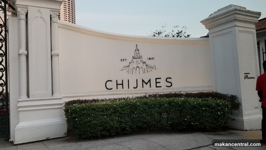 White wall with 'CHIJMES' logo and 'EST. 1841' above a stylized building illustration; flanked by classical white pillars; low green hedge in front; paved ground; partial view of person in red shirt on right.