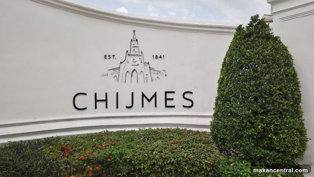 White curved wall with black embossed logo featuring a stylized building and "EST. 1841" above the name "CHIJMES" in bold letters. Lush green shrubbery and flowering plants at base, with a manicured conical tree to right. Sky visible above.