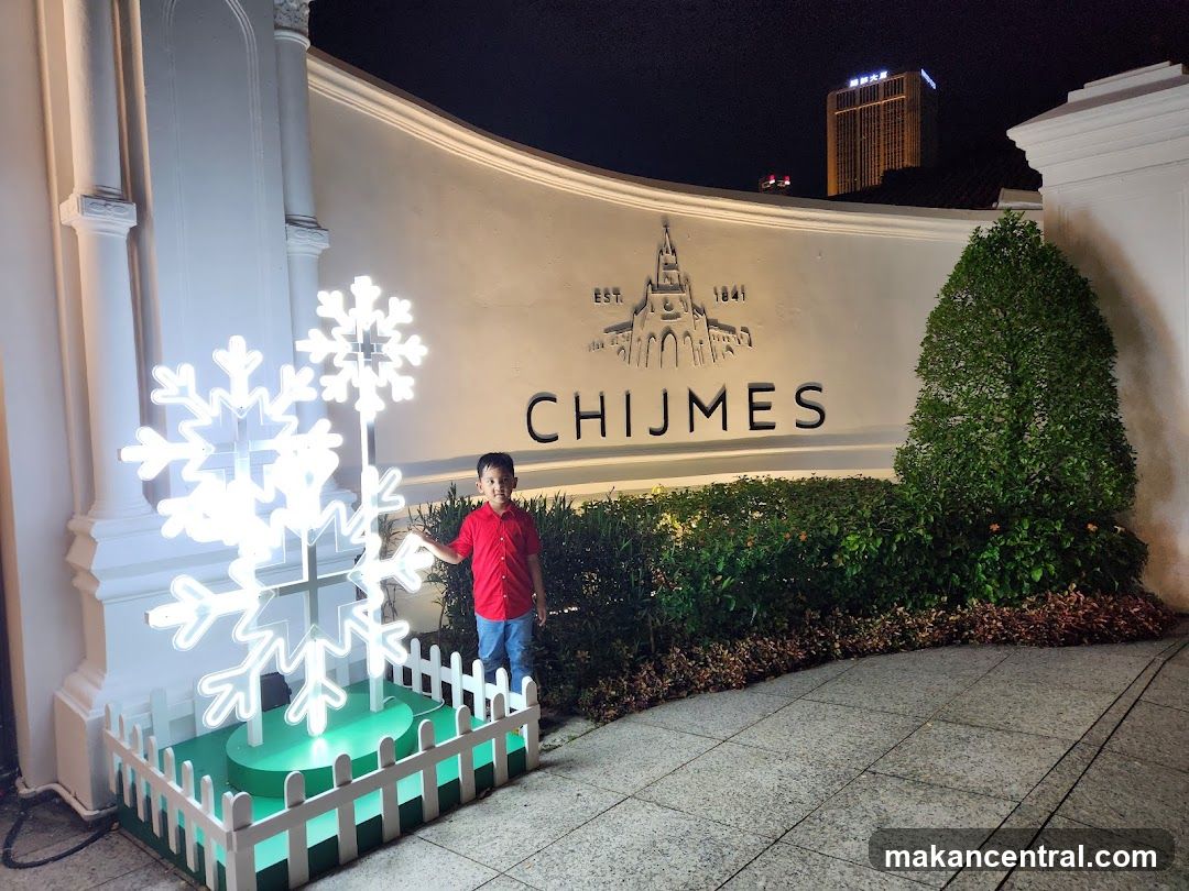 Nighttime exterior shot of Chijmes with illuminated signage and decorative snowflake display. A child in a red shirt stands beside the display. The building facade features 'EST. 1841' and 'CHIJMES' engraved on a curved wall, flanked by manicured hedges and classical architectural elements. In the background, a tall lit building is visible against the dark sky.