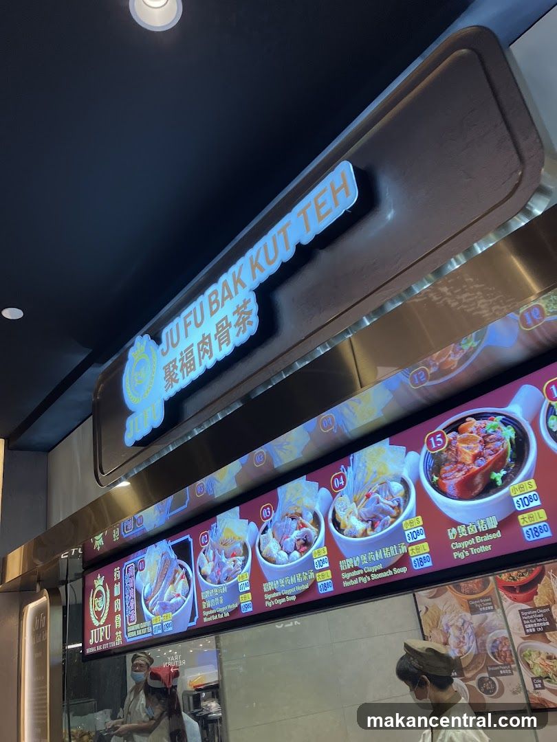 Storefront with illuminated sign displaying 'JU FU BAK KUT TEH' and Chinese characters '聚福肉骨茶'. Below the sign is a digital menu board showing food images and prices in Singapore dollars. Two staff members wearing masks are partially visible inside the shop.