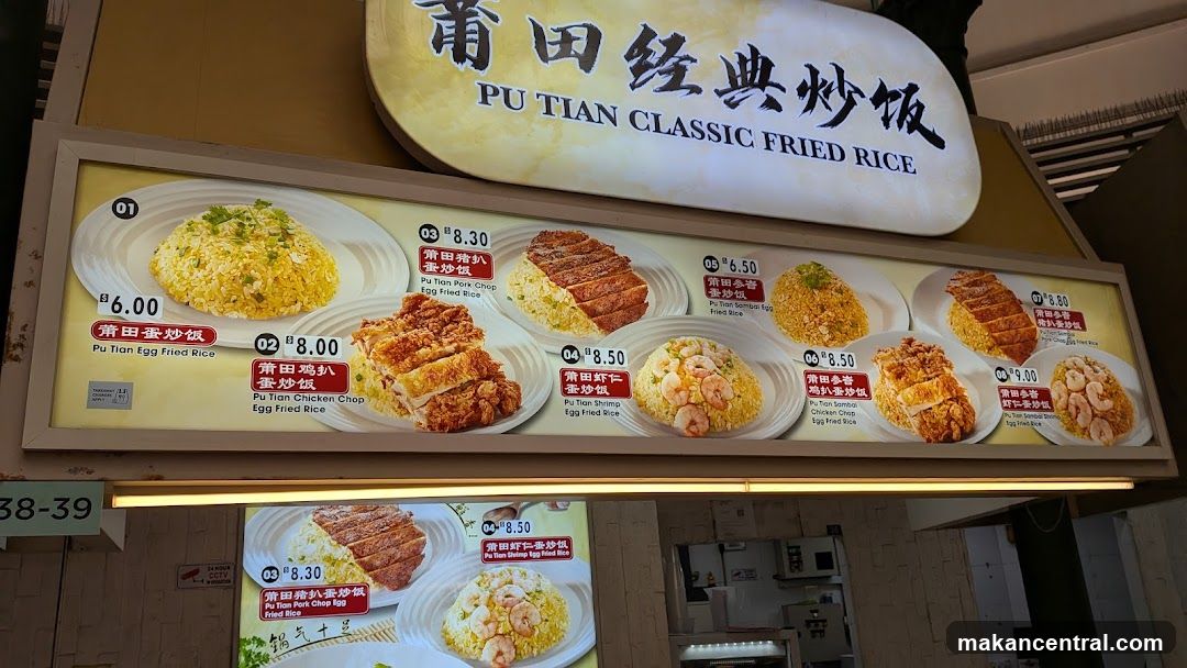 A large illuminated menu board displaying various fried rice dishes with prices in Singapore dollars. The sign above reads 'PU TIAN CLASSIC FRIED RICE' in both Chinese and English. Multiple dish options are shown with images, including Pu Tian Egg Fried Rice, Pu Tian Chicken Chop Egg Fried Rice, Pu Tian Pork Chop Egg Fried Rice, etc., each with a price tag.