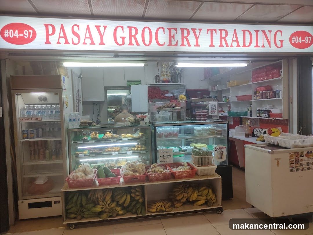 Storefront with red lettering on white sign above entrance, unit #04-97. Visible interior includes refrigerated display cases, shelves stocked with goods, and a counter with fresh produce like bananas displayed in baskets. A staff member is partially visible behind the counter.