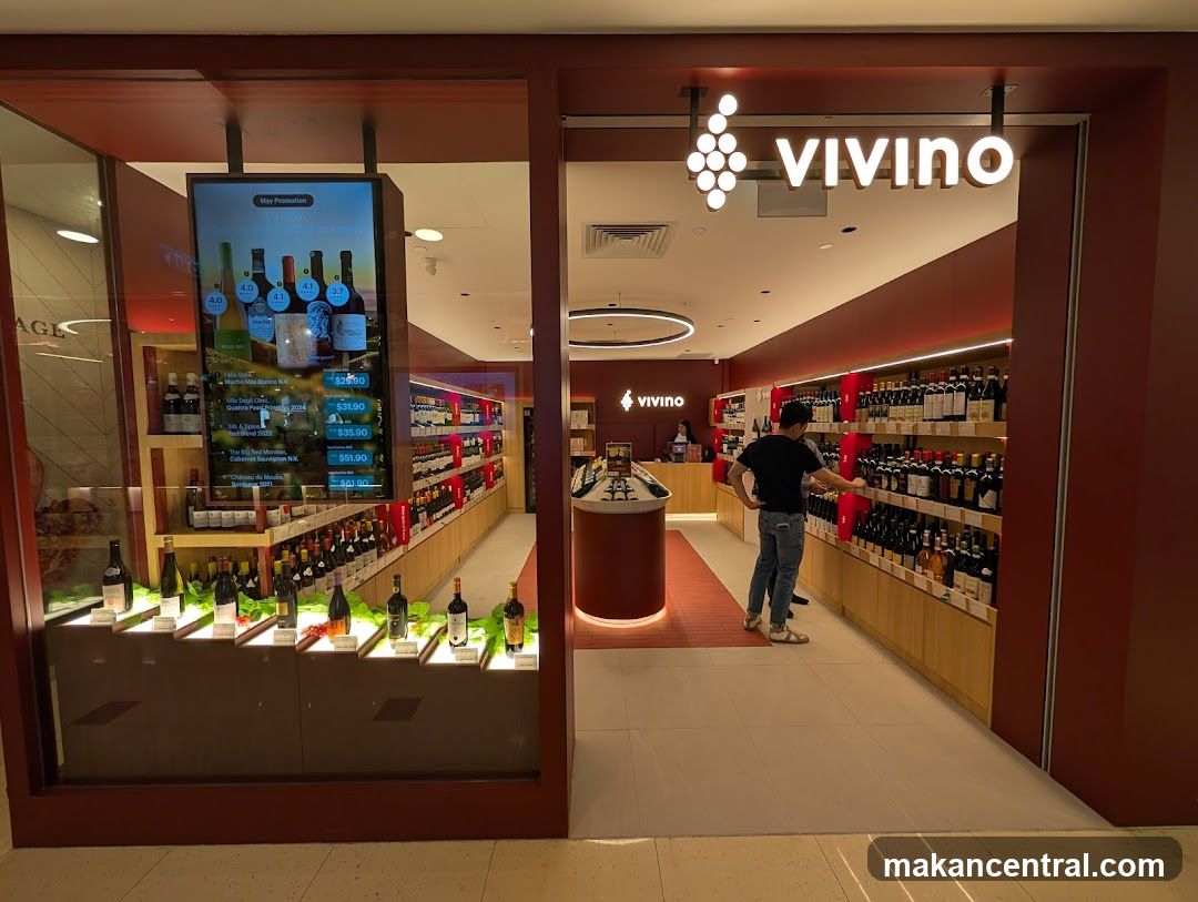 Storefront with red-toned facade, illuminated 'vivino' logo above entrance. Glass window displays wine bottles on lit shelves and a digital screen showing promotions. Interior view shows customer browsing racks of wine, circular counter, and ambient lighting.