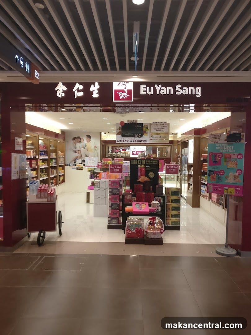 Store entrance with illuminated sign displaying "余仁生" and "Eu Yan Sang" in white lettering on a dark red background. The ceiling features a striped pattern of black and silver lines. Inside the store, shelves are stocked with products, promotional displays are set up near the entrance, and various sale signs and advertisements are visible. A small cart is positioned to the left of the entrance.