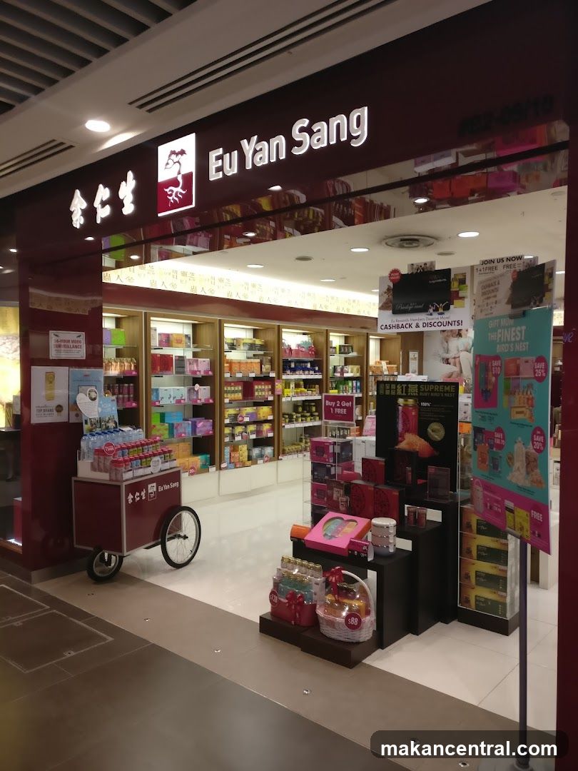 Store entrance with illuminated sign displaying 'Eu Yan Sang' in English and Chinese characters above the glass facade. Interior shelves stocked with products are visible through the storefront. A red promotional cart is parked outside the entrance. The ceiling has recessed lighting, and a display stand with promotional materials stands near the entrance.