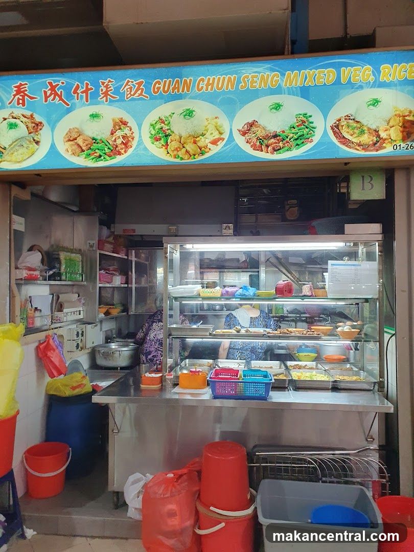 Exterior view of a food stall with a blue signboard displaying the name 'Guan Chun Seng Mixed Veg. Rice' in English and Chinese characters. The sign features images of four different rice dishes. Below, a glass display case holds various prepared foods. Inside, staff are visible working behind the counter. The shop has stainless steel counters, red plastic buckets, and shelves stocked with ingredients. A small green 'B' sign is mounted on the right side.